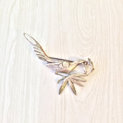 Vintage sterling silver bird brooch designed by Guglielmo Cini for Gump's, featuring a detailed bird with outstretched wings perched on a branch, crafted in a mix of polished and textured silver against an off-white woodgrain background.