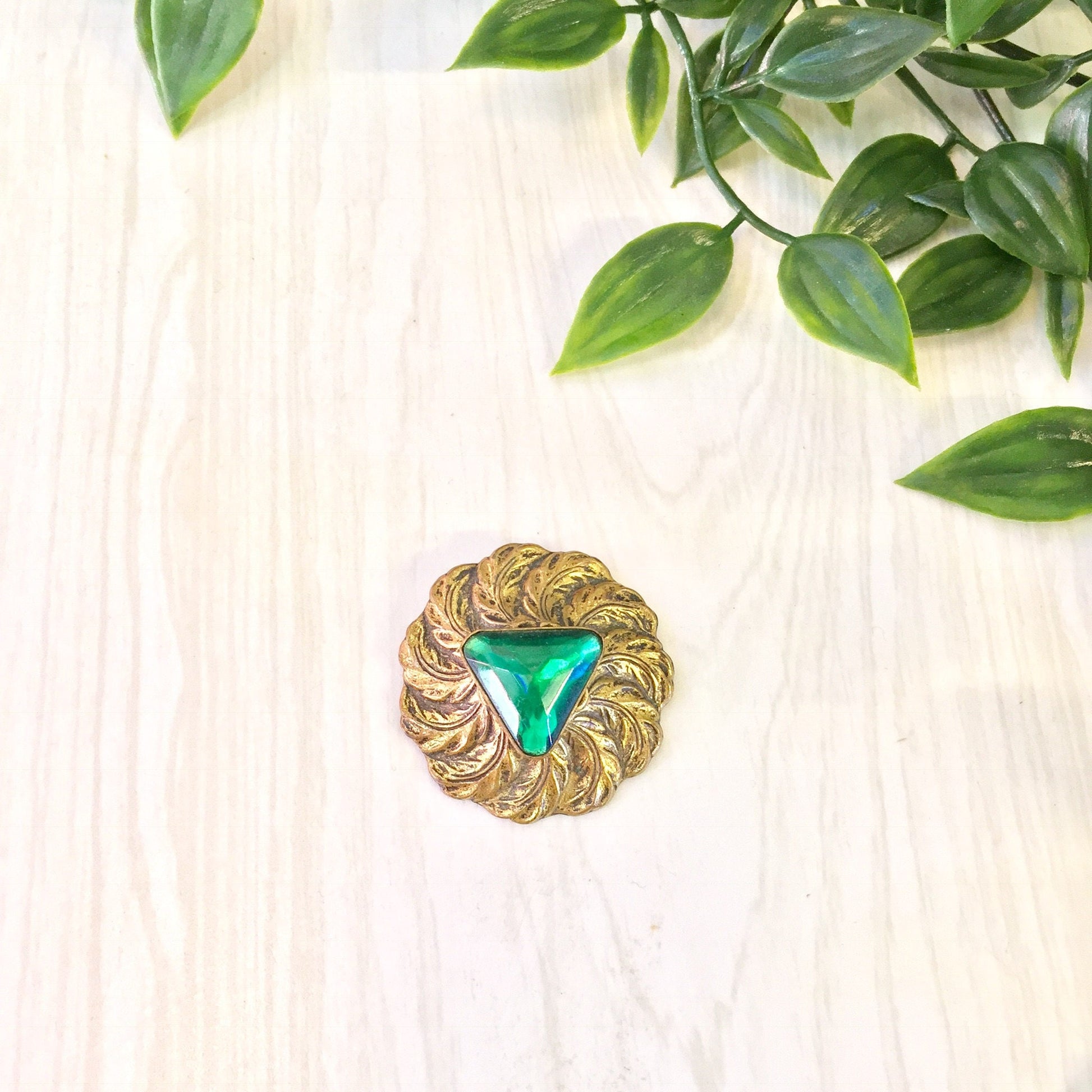 Vintage gold tone round brooch with green center stone surrounded by feather-like design, displayed on light wooden surface with green leaves in background.