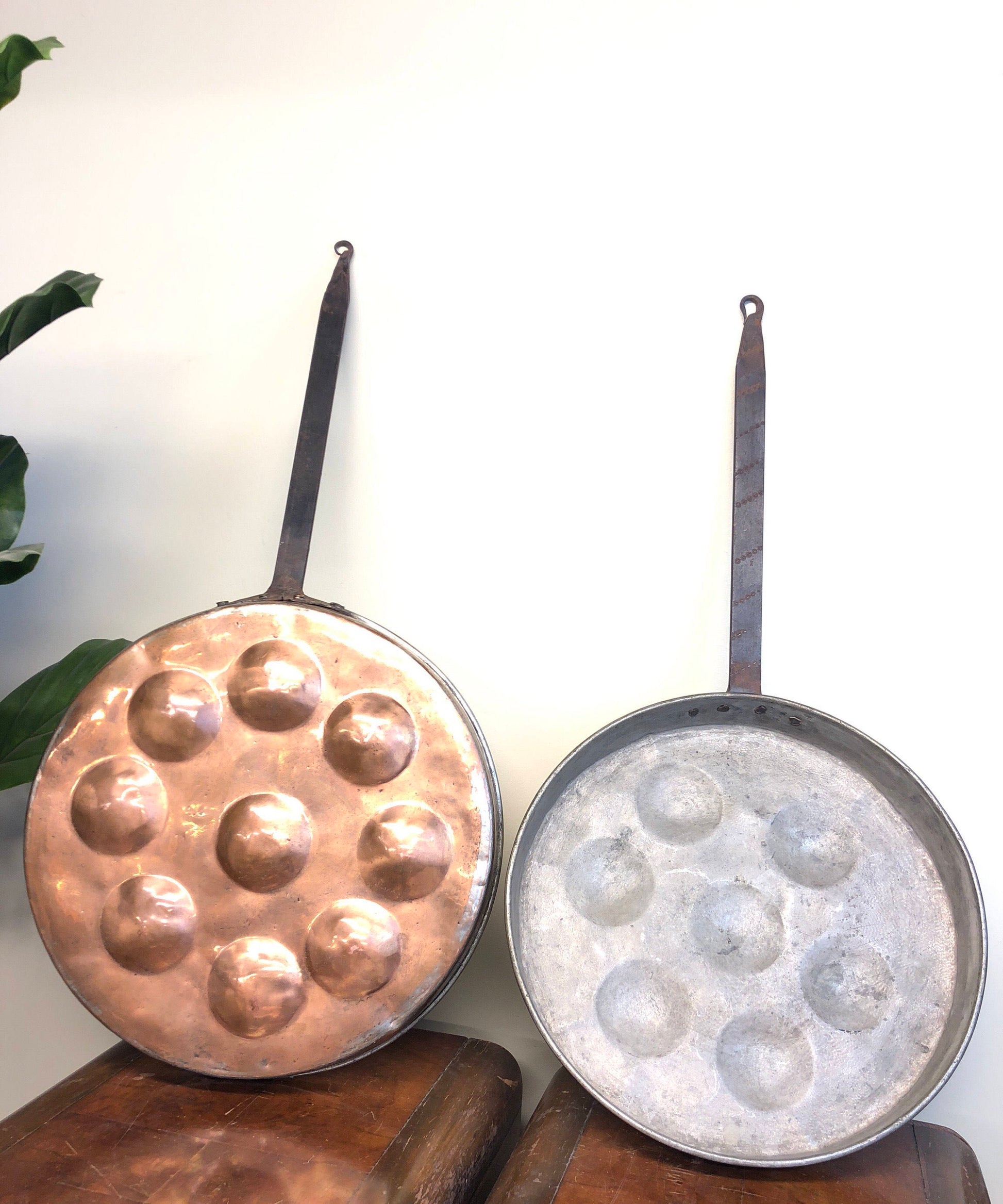 Vintage copper and aluminum frying pans with dimple textures for poaching eggs, rustic hand-crafted kitchenware decor set on wooden surface with plant leaves in background