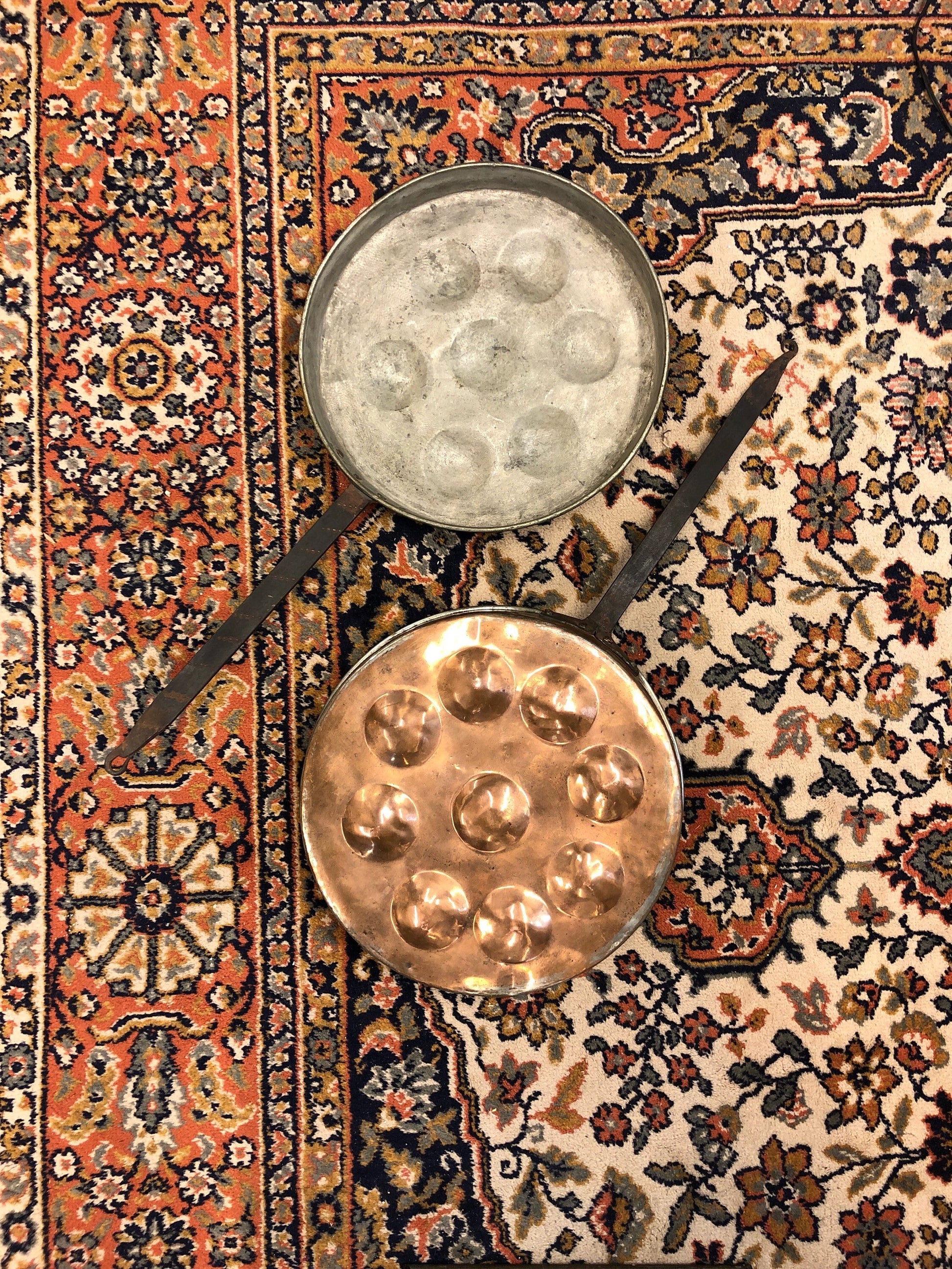 Antique copper pans and ladle on vintage patterned rug. Round pans with patina and dimpled texture for poaching eggs, displayed on colorful oriental carpet with floral and geometric designs in orange, navy blue and cream colors. Rustic folk art kitchenware and home decor.