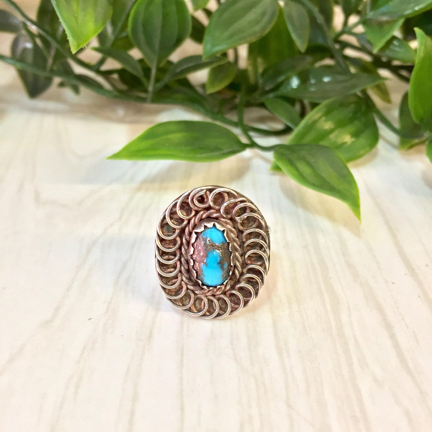 Vintage sterling silver ring with oval turquoise stone set in intricate swirled design, placed on light wooden surface with green leafy plant in background.