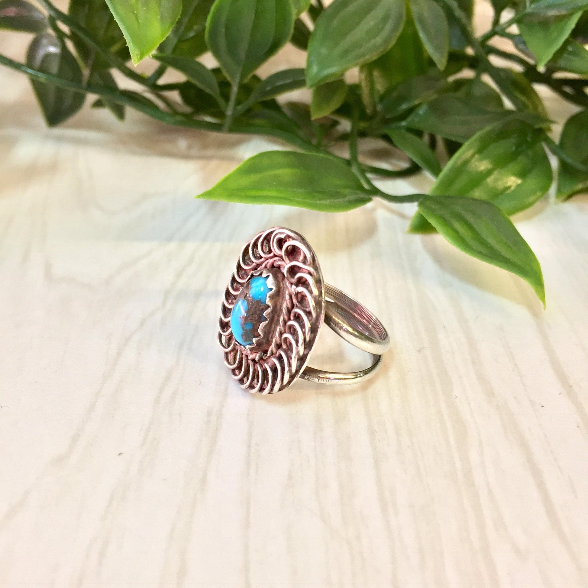 Vintage silver and turquoise statement ring with ornate filigree design, set against wood surface with green foliage in background.