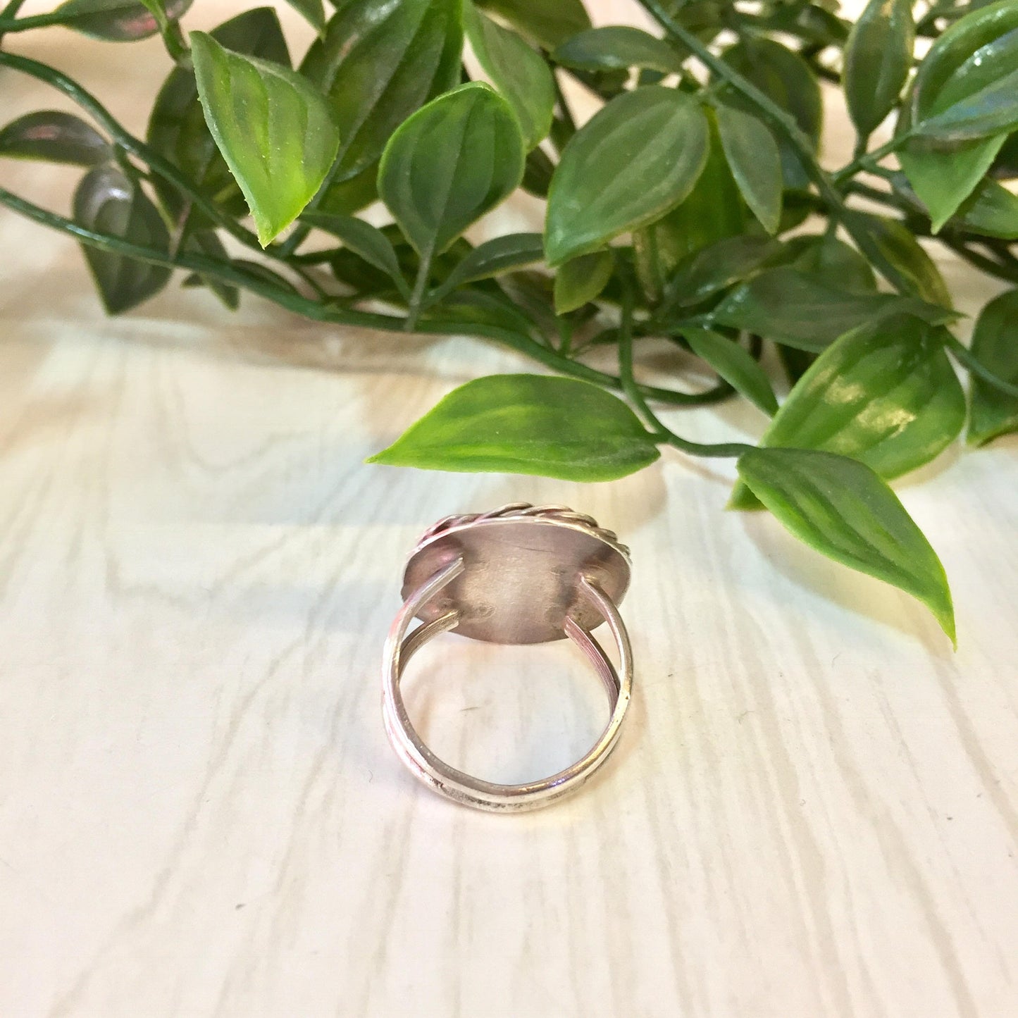 Vintage sterling silver ring with an oval turquoise stone set against green leafy plant stems on a light wood background.