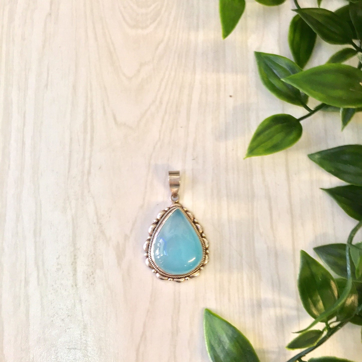 Vintage silver and opalite pendant necklace with blue and green hues on a wooden surface surrounded by green leaves.