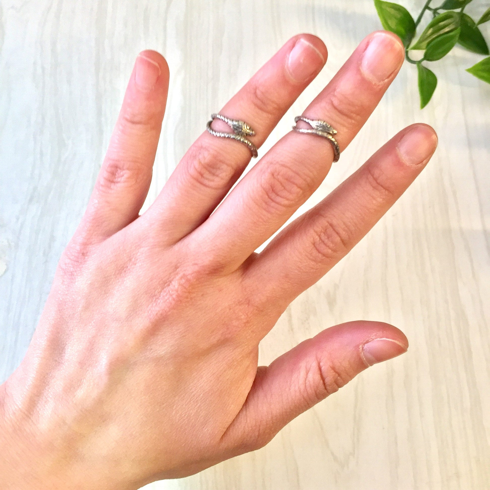 Silver snake rings worn on fingers against light wood background with green leaves