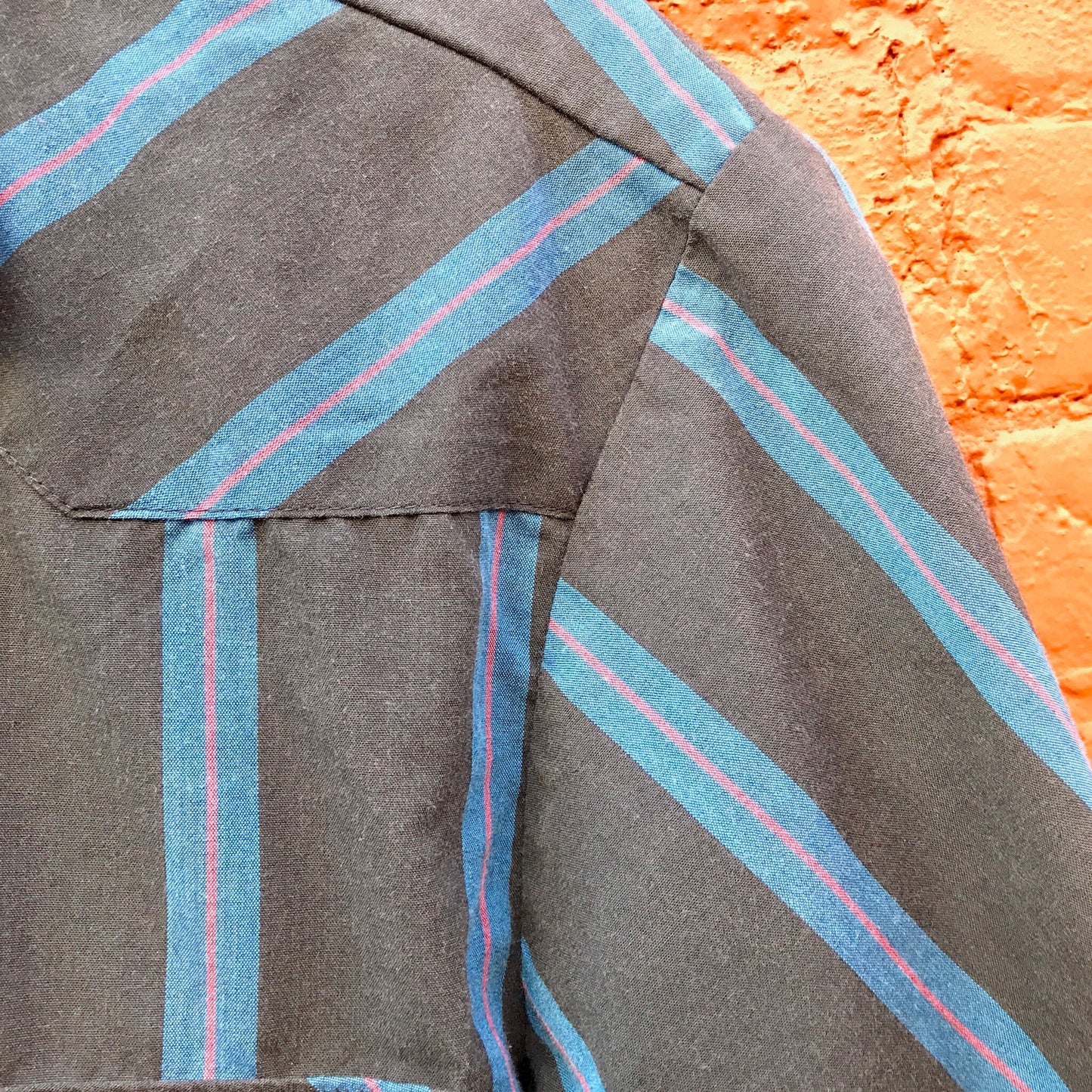 Vintage striped short-sleeved button-down Wrangler western shirt with blue and gray stripes, close-up detail of collar and shoulder area, paired with brick wall background.