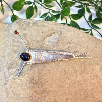 Vintage silver abstract brooch with geometric design, featuring a black onyx stone and red accents, resting on a textured stone surface with green leaves in the background.