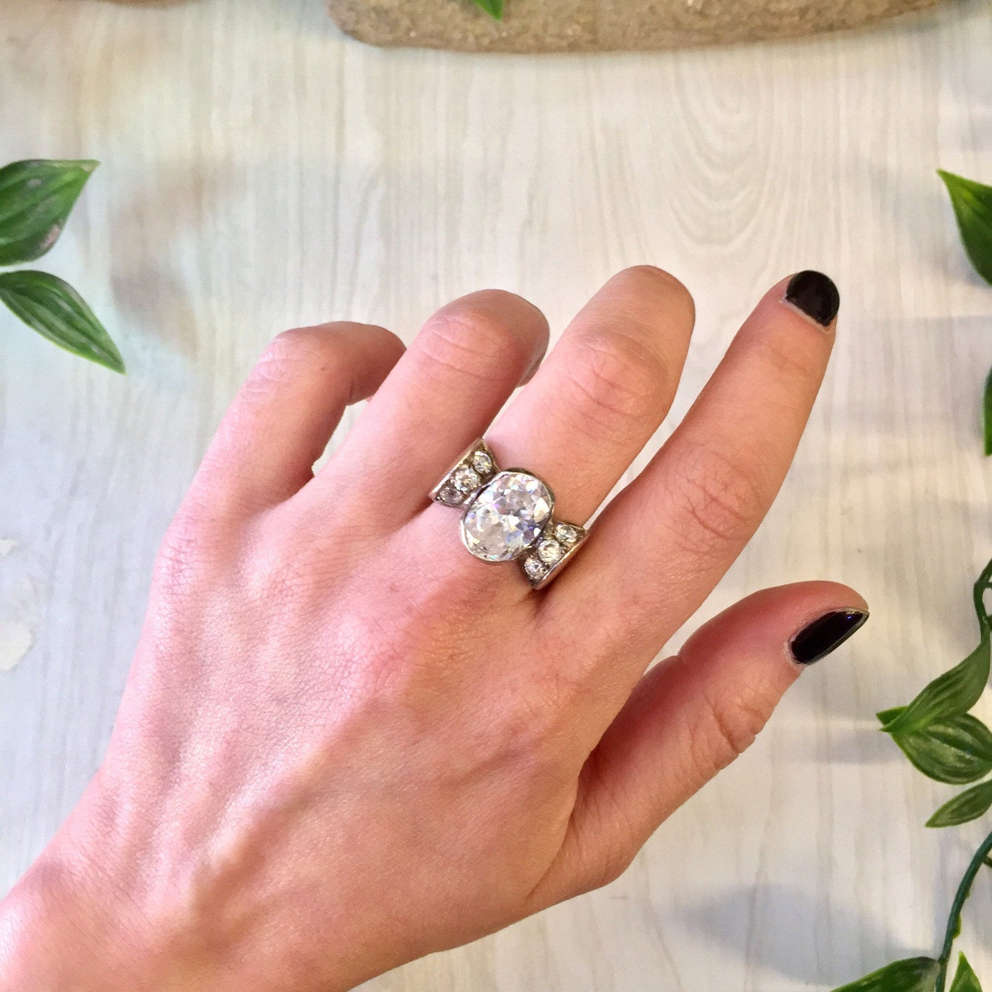 Vintage sterling silver statement ring with sparkling round diamond on a hand, surrounded by green leaves on a light wooden surface.