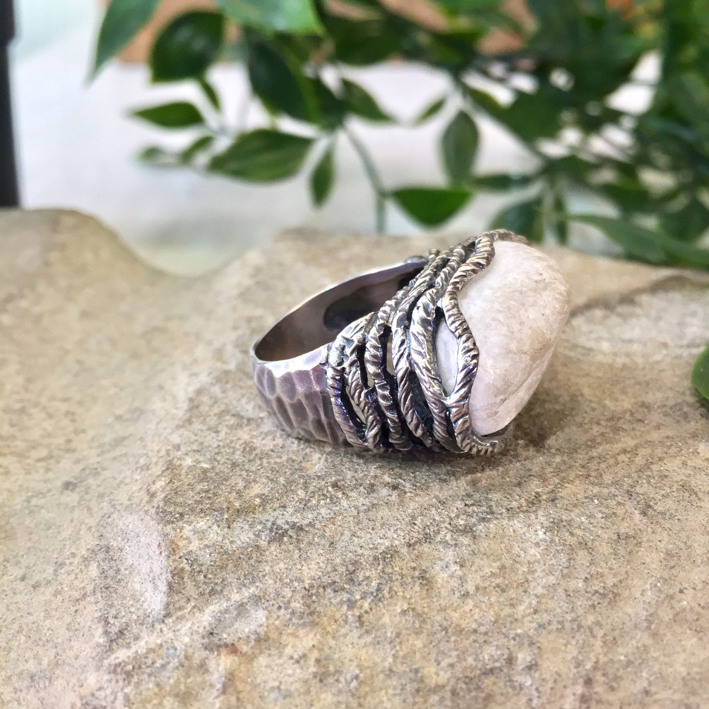 Vintage sterling silver ring with hammered texture and twisted rope setting holding a large quartz stone, sitting on natural rock surface with green foliage in soft focus background.