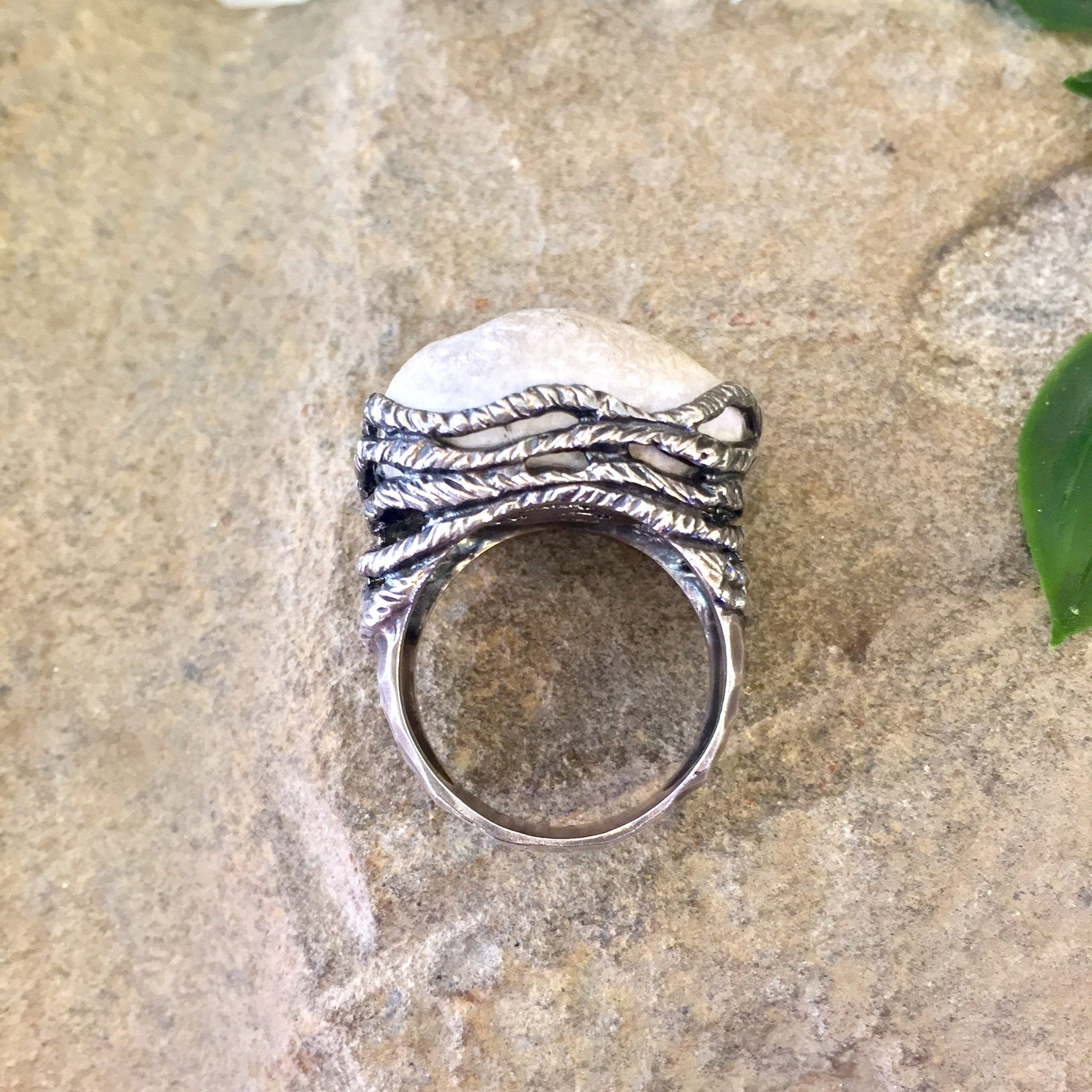 Vintage sterling silver ring with rope-textured band and large quartz stone set in hammered silver prongs, placed on weathered stone surface with green leaves in background.