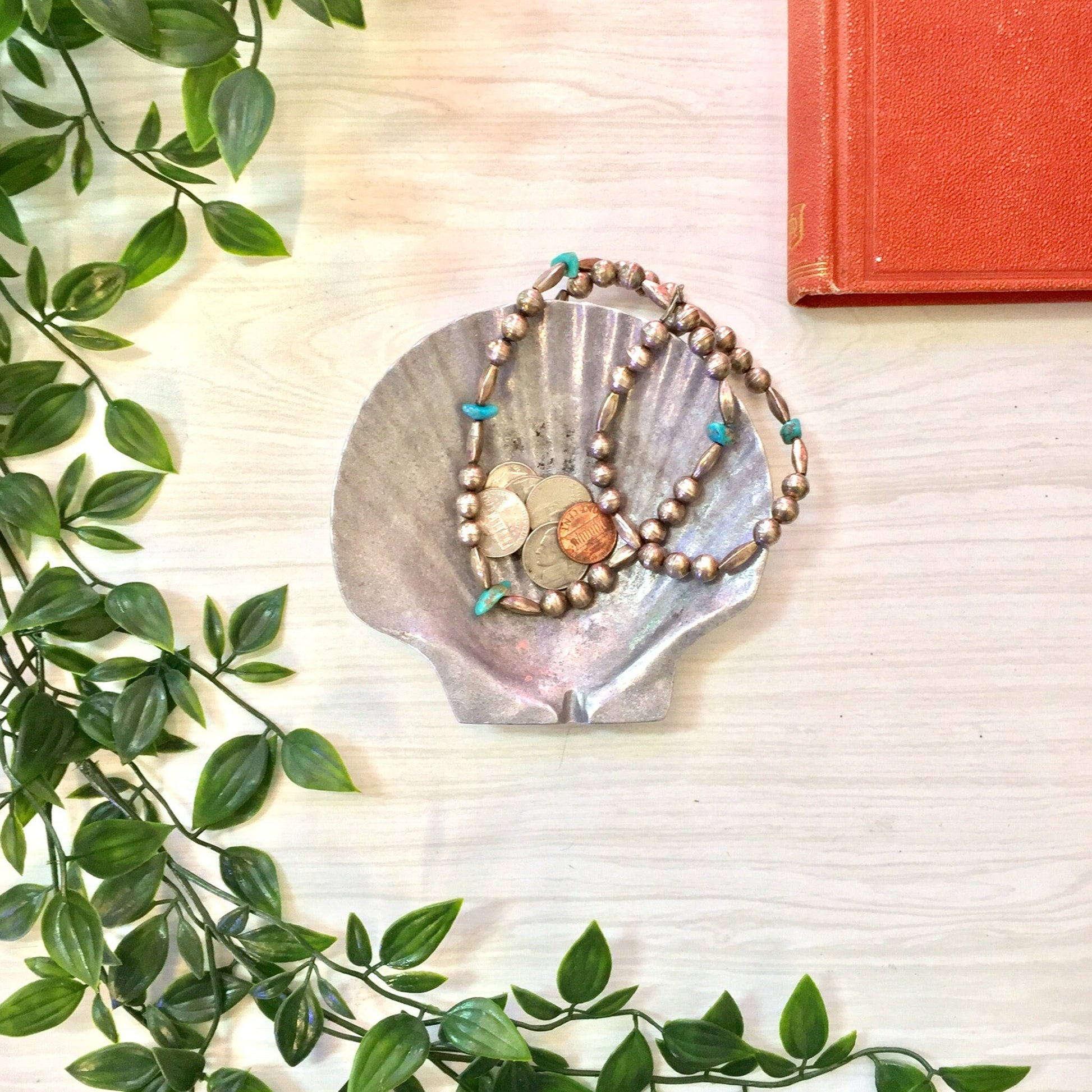 Vintage pewter seashell-shaped ashtray or trinket dish adorned with colorful stones, displayed on a white wooden surface with green foliage and a red book.