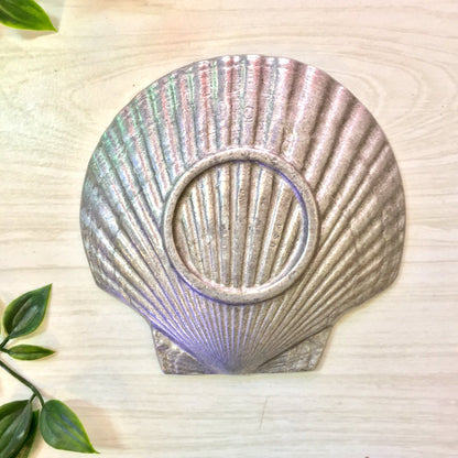 Vintage pewter seashell-shaped ash tray or catch-all dish with iridescent purple and silver tones, displayed on a light wooden surface with green leaves.