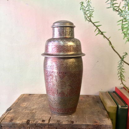 Vintage etched and painted metal Indian cocktail shaker with silver and red floral design, placed on a wooden surface next to books.