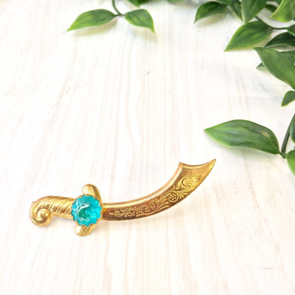 Vintage gold tone scimitar brooch pin with turquoise gemstone accent, displayed on light wood surface with green foliage in the background.