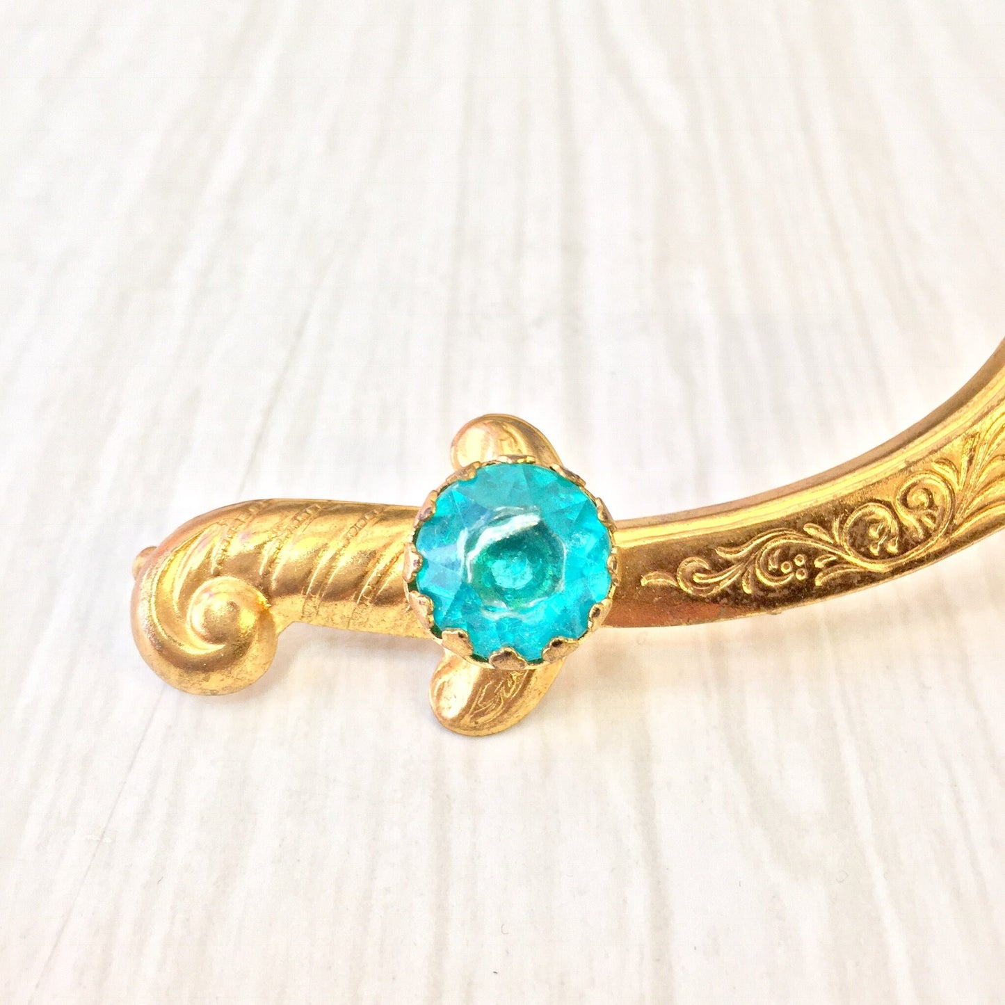 Vintage gold-tone sword brooch with teal gemstone accent, featuring intricate scroll designs on the handle and blade of the scimitar-shaped pin.