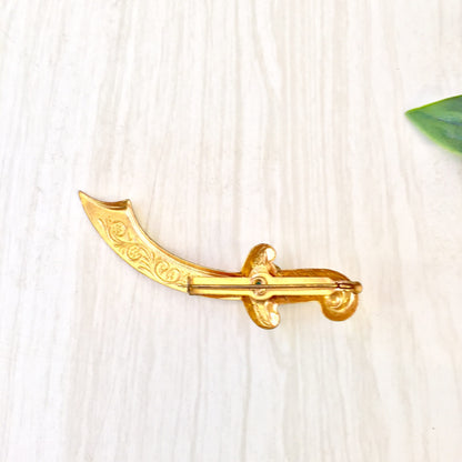 Vintage gold-tone sword brooch with decorative scroll design on the blade, resembling a scimitar, pinned to a light wooden surface with a green leaf in the background.