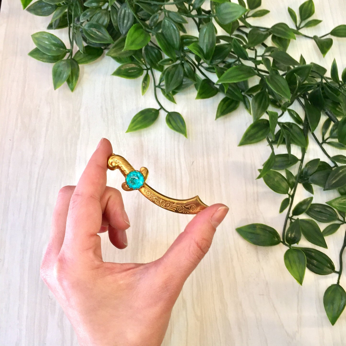 Hand holding a gold-tone vintage sword brooch with a turquoise gemstone against a background of green leaves