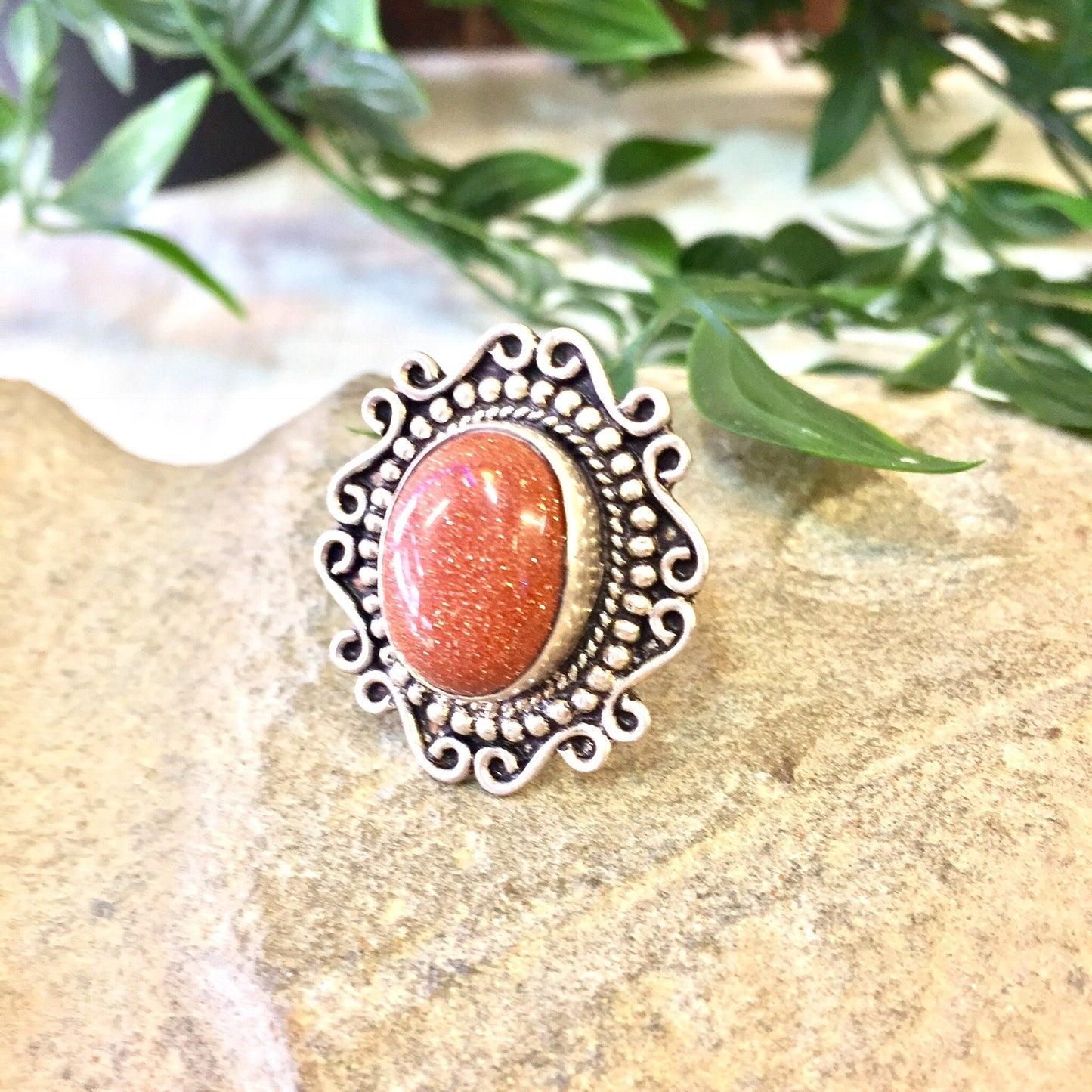 Vintage sterling silver ring with orange druzy quartz stone and intricate Celtic design border, displayed on rough beige stone surface with green leafy plants in soft focus background.