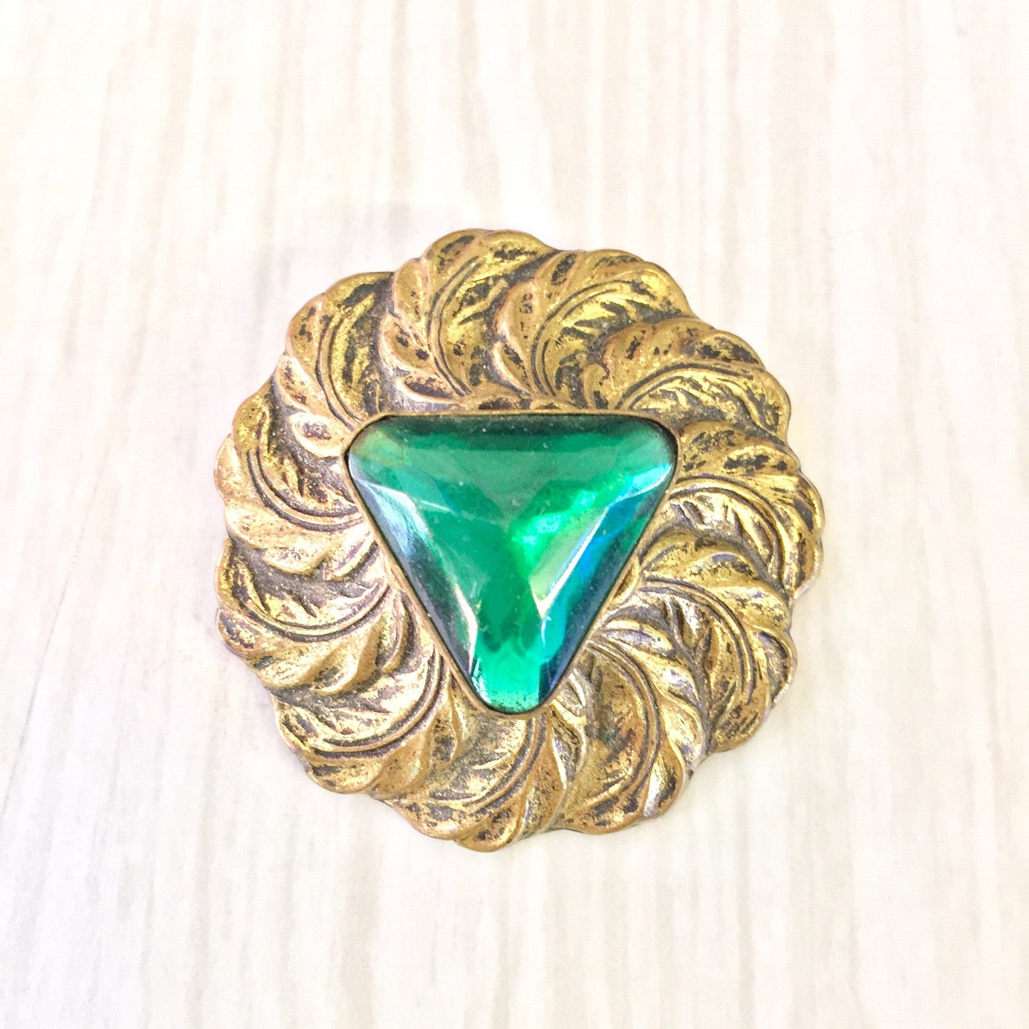 Vintage gold tone round brooch with green triangular center stone surrounded by textured feather-like design, costume jewelry pin for Valentine's Day or any occasion.