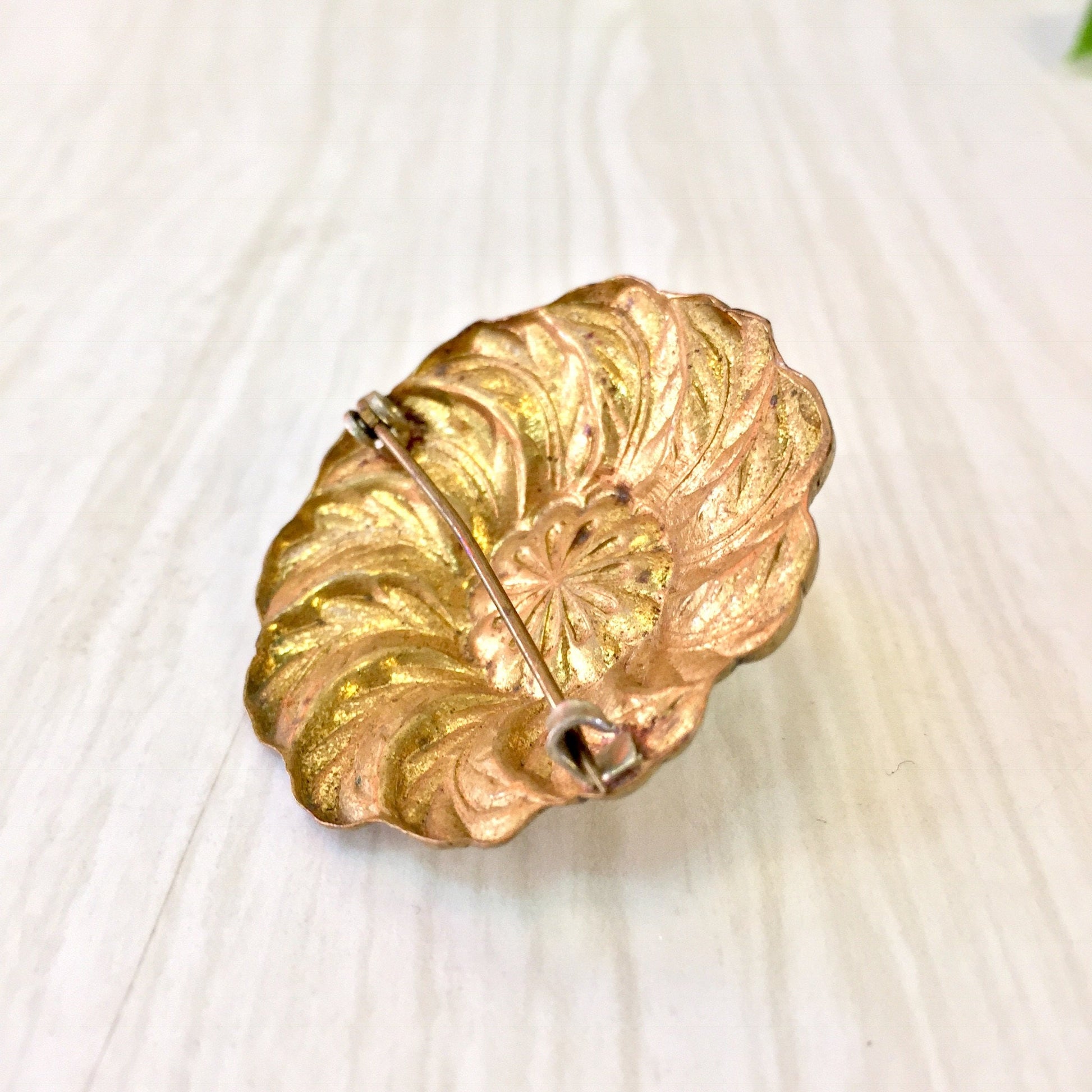 Vintage gold tone leaf-shaped brooch pin with detailed embossed pattern, resembling a curled feather or leaf, on a light wooden surface background.