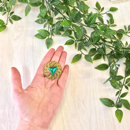 Green and gold vintage round feather brooch pin held in hand over white fabric with greenery