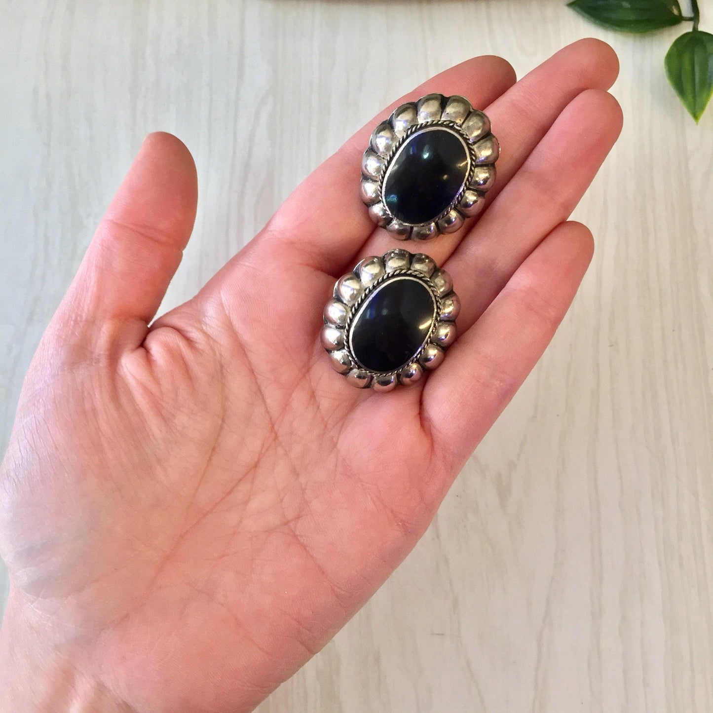 Vintage sterling silver clip-on earrings with black onyx stones in a Southwestern style, held in a person's hand against a light wooden background with leaves.