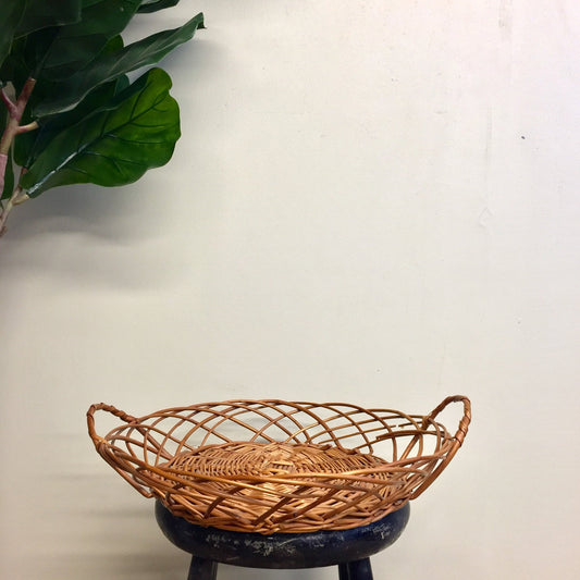 Vintage woven rattan basket with handles on wooden stool, green plant leaves in background, bohemian home decor