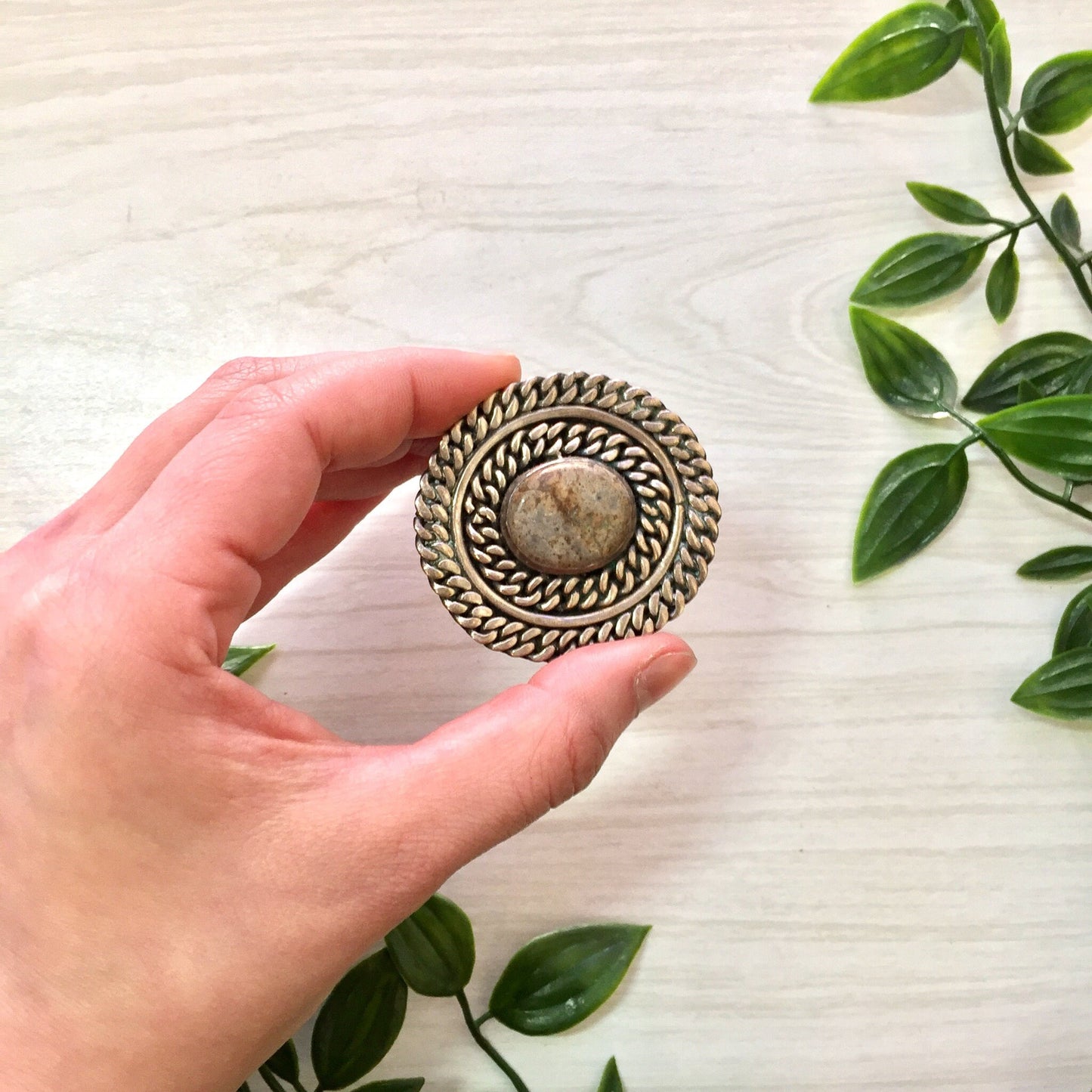 Vintage sterling silver round brooch with natural stone from Taxco, Mexico, held in hand near green plant leaves on white wooden surface, perfect gift for mom or vintage jewelry collector.