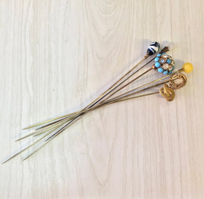 Vintage hat pins with colorful beads and decorative tops, including blue, yellow, black and white designs, arranged on a light wooden surface.