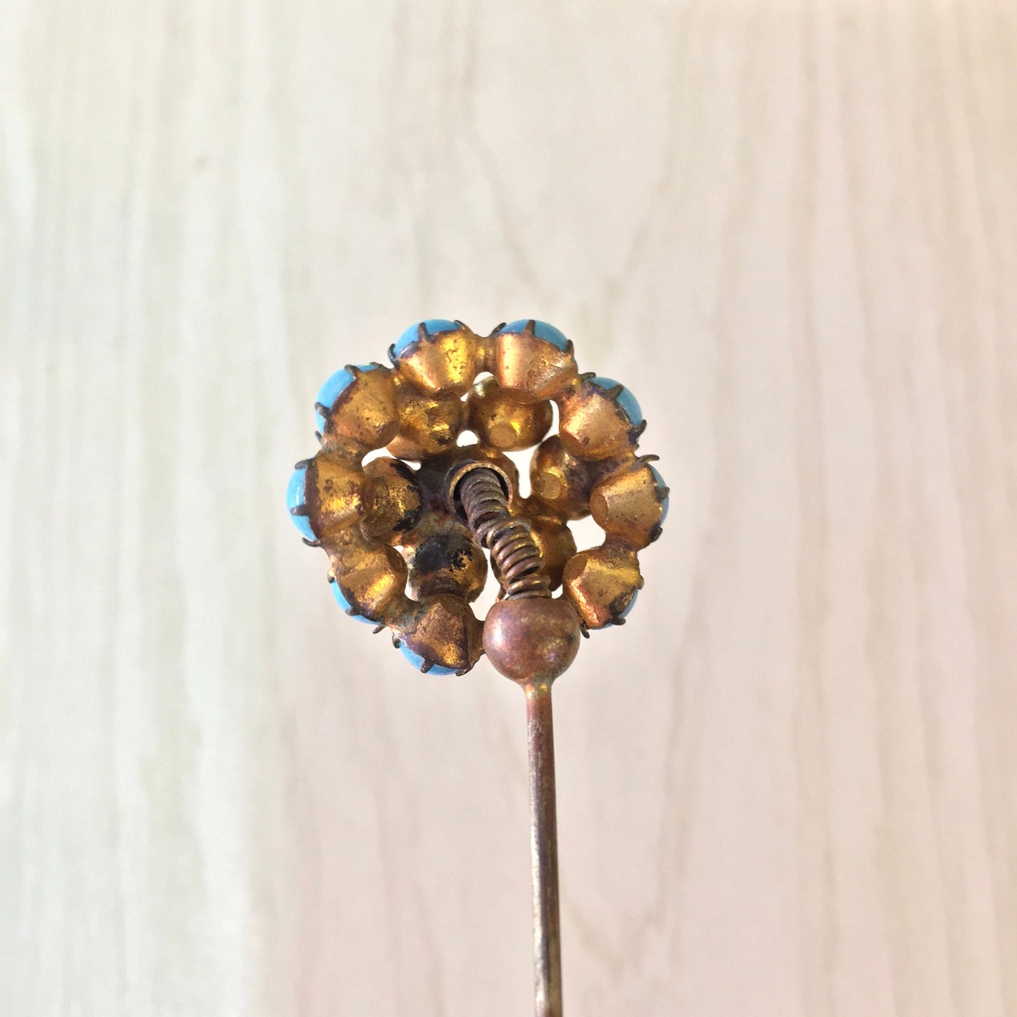 Vintage golden-colored hat pin with blue accents resembling a flower, shown against a neutral fabric background.