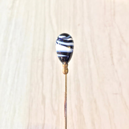 Alt text: Vintage blue and white swirl pattern glass hat pin with brass shaft on light wood background