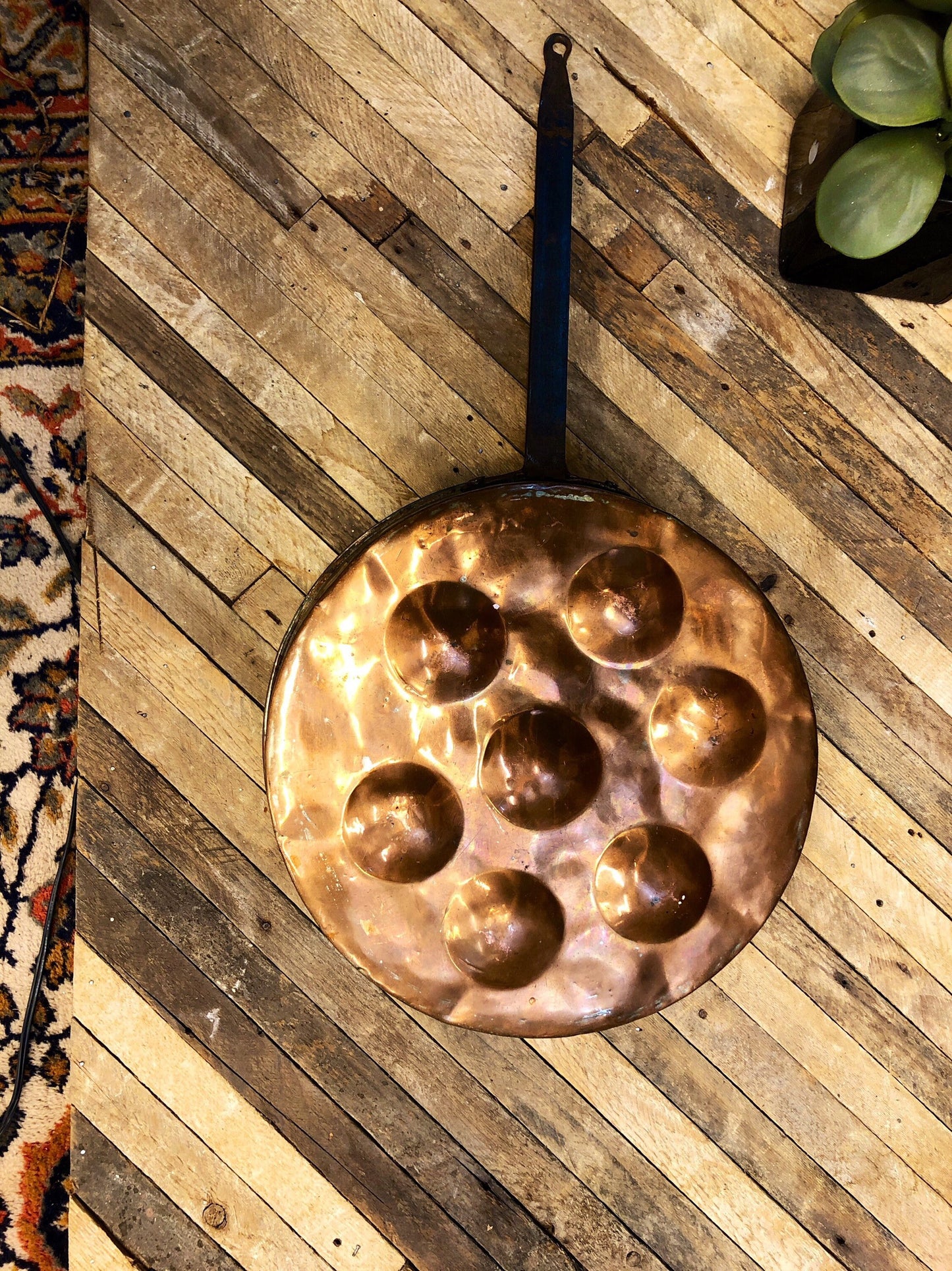 Vintage copper pan with seven dents for poaching eggs on rustic wooden surface, alongside ornate rug and green apples