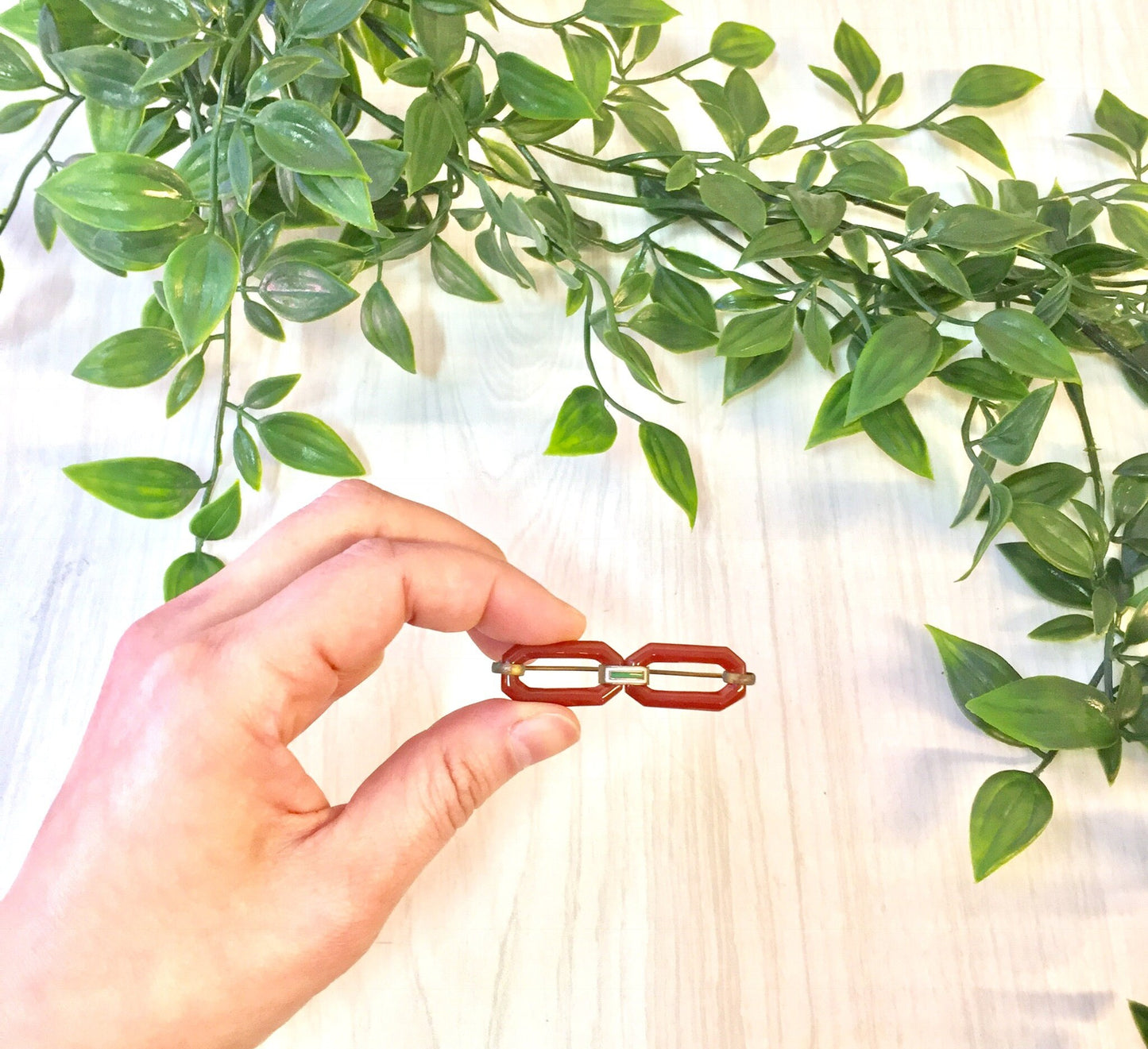 Woman's hand holding a vintage red brooch made of carnelian against a background of green leaves on a white cloth surface.