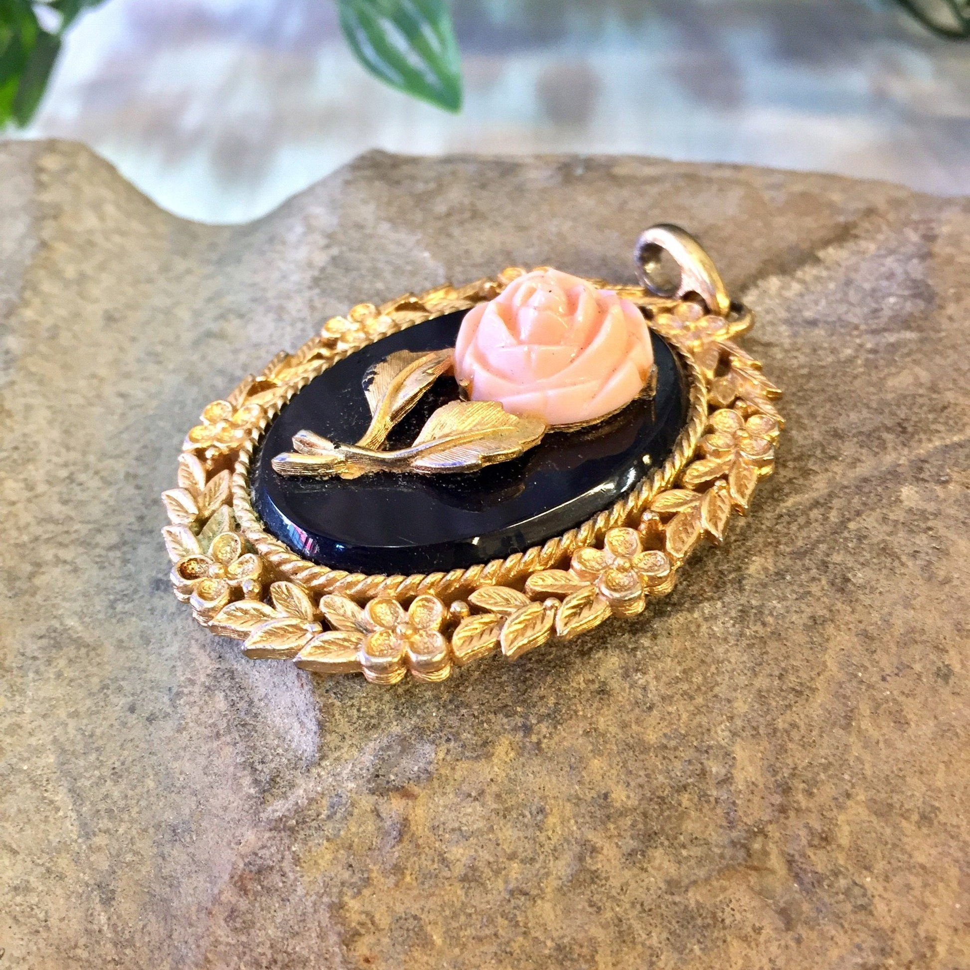 Vintage gold-toned cameo pendant featuring a pink rose on a black oval background, surrounded by an intricate leaf design border, displayed on a rock surface.