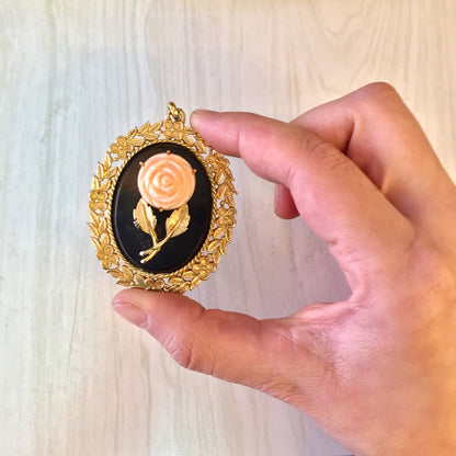 Vintage gold-toned cameo pendant featuring an intricately carved pink rose against a black background, held in a person's hand to showcase the details and size of the elegant Avon mirror jewelry piece.