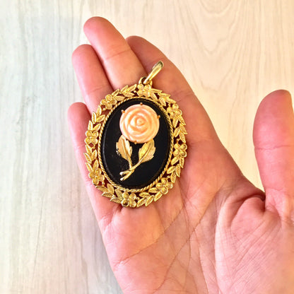 Vintage gold-toned cameo pendant featuring a delicate pink rose on a black background, held in a person's hand. The pendant has an ornate gold frame with leaf and vine detailing surrounding the oval cameo.