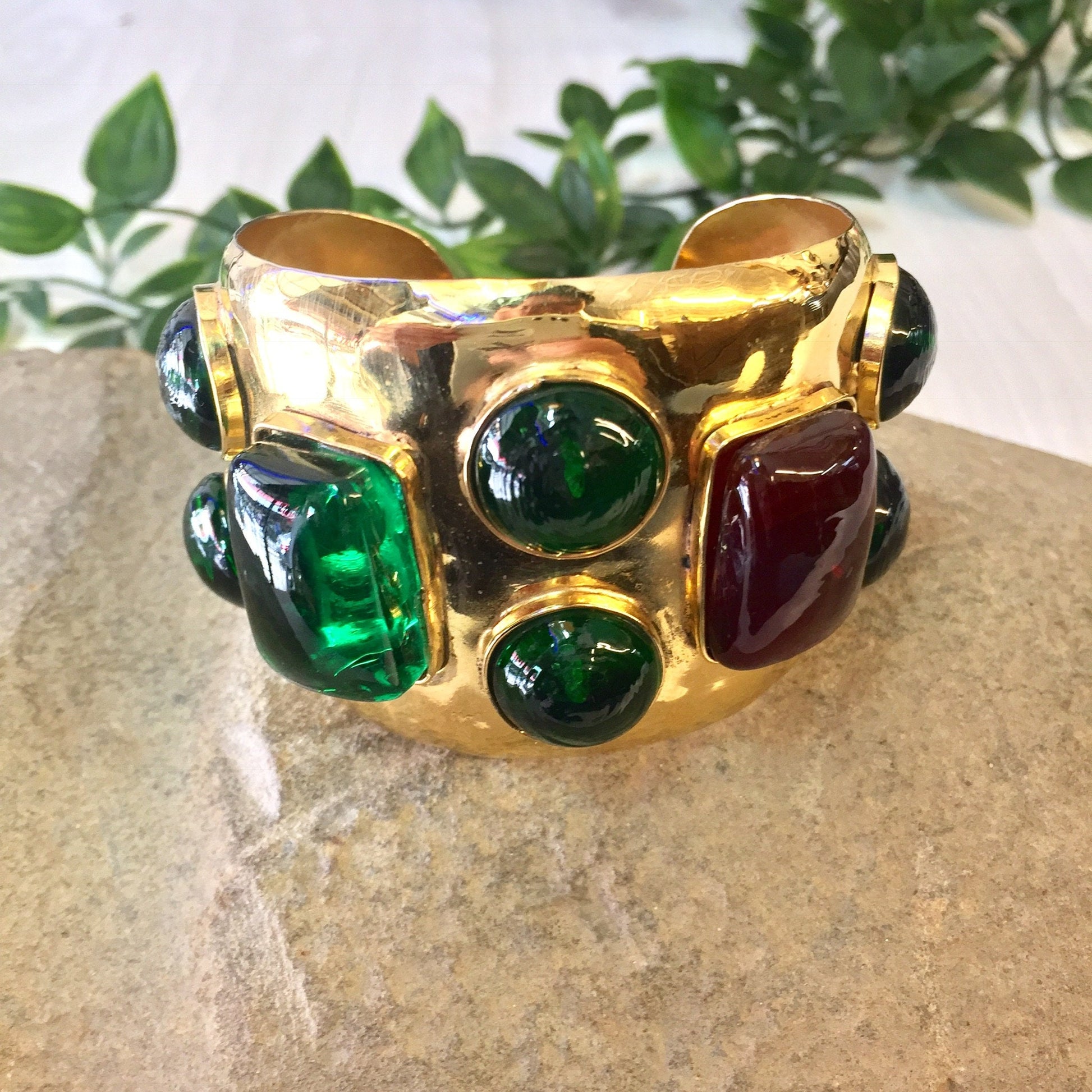 Vintage gold-toned cuff bracelet featuring large green and red glass beads set in ornate metalwork on a textured stone surface with greenery in the background.