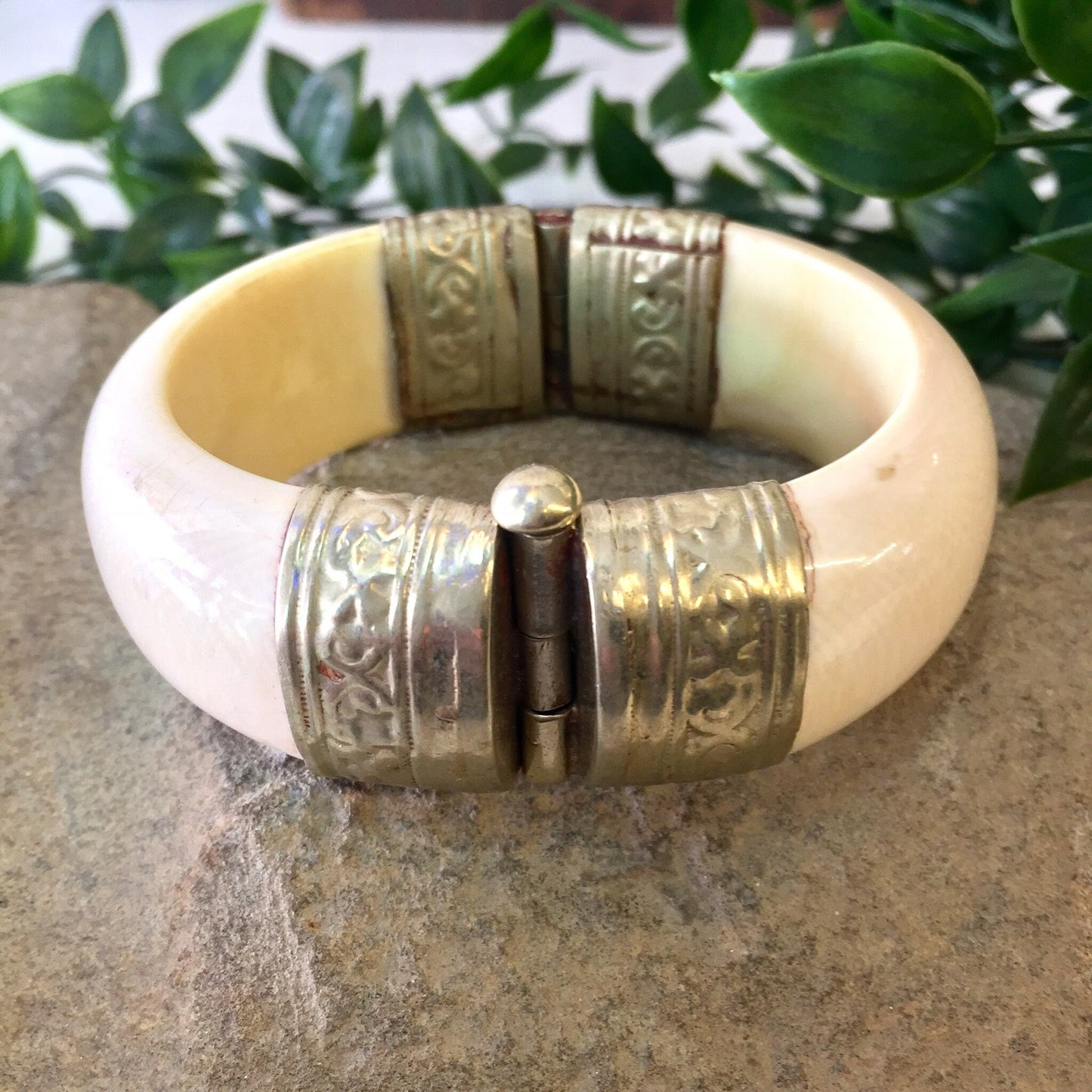 Vintage white and silver hinged bangle bracelet with engraved designs and pin closure, displayed on stone surface with greenery in background.