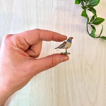 Two small decorative bird figurines resting on a person's finger in front of a wooden background with a plant in view.