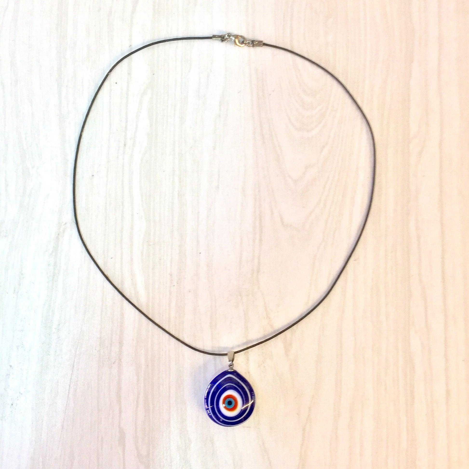 Evil eye pendant necklace featuring blue, red and white concentric circles on a translucent glass teardrop-shaped charm hanging from a thin metal chain against a light wood background.