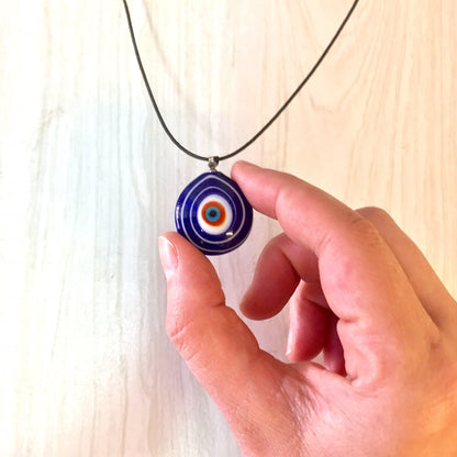 Vintage glass evil eye pendant necklace held in hand, featuring concentric blue, white, and red circles resembling an eye, on a black cord against a light wooden background.