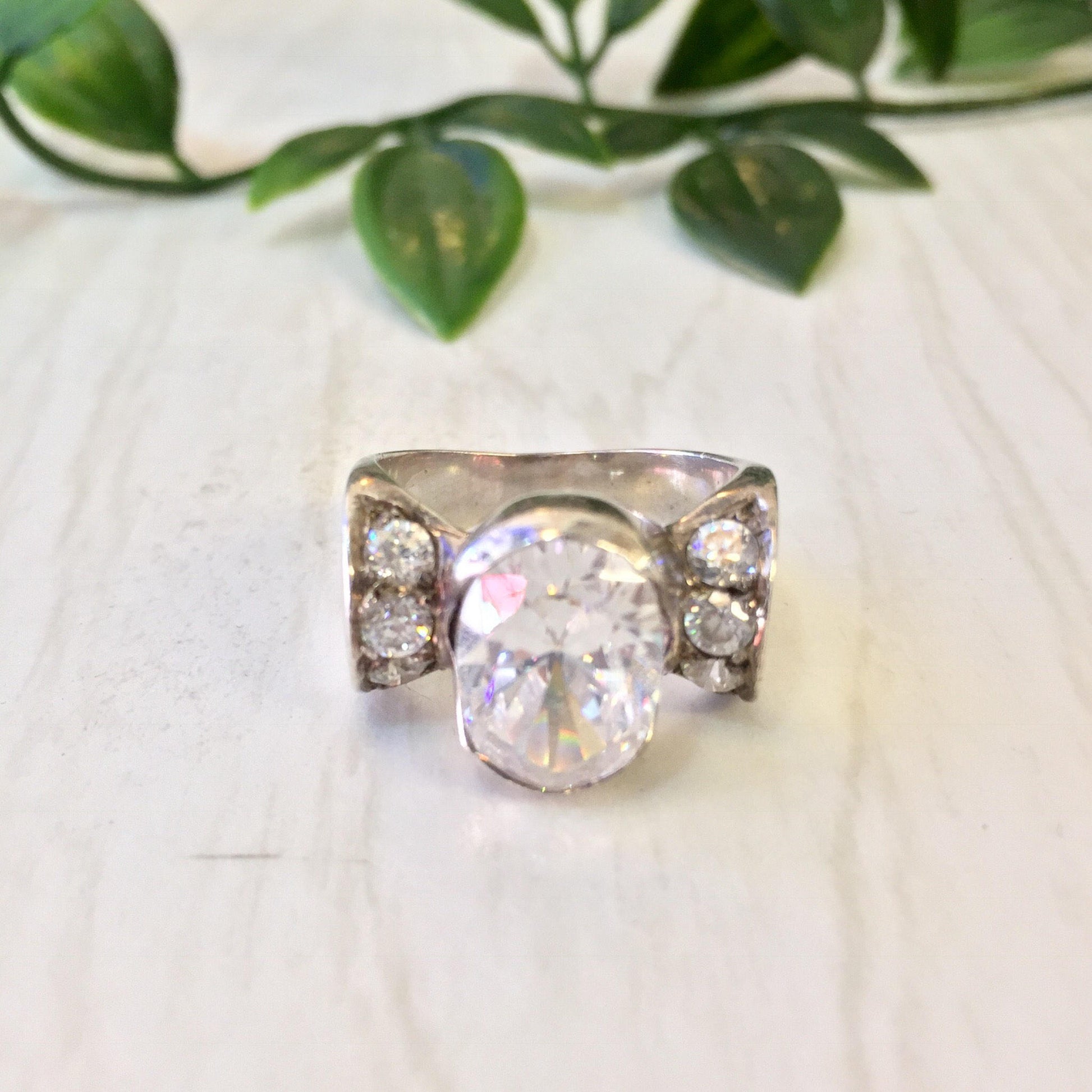 Vintage sterling silver engagement ring with large central stone and accent diamonds on the sides, displayed on a white background with greenery in the background.