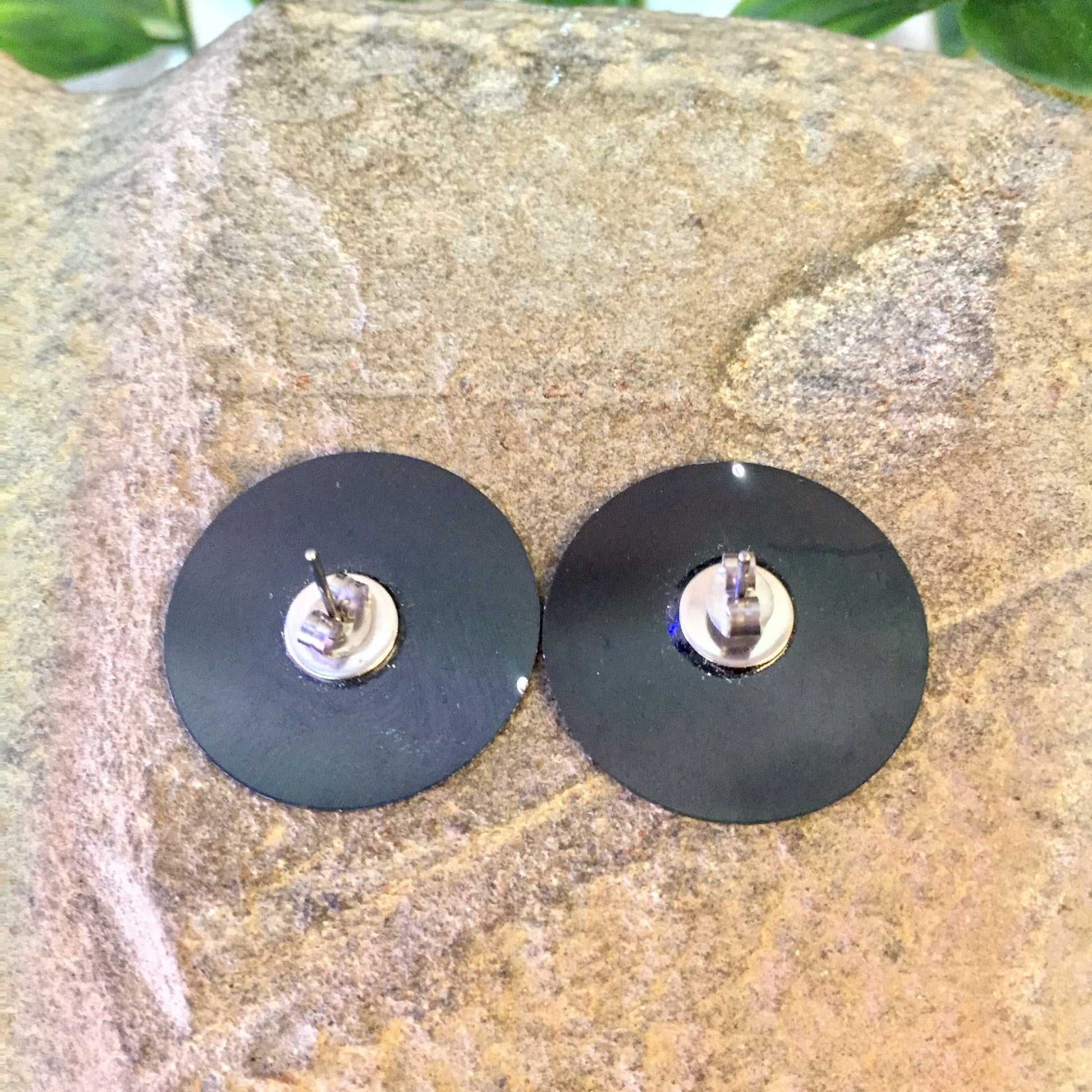 Vintage black disc earrings with small clock face design, displayed on stone surface with leaves in background.