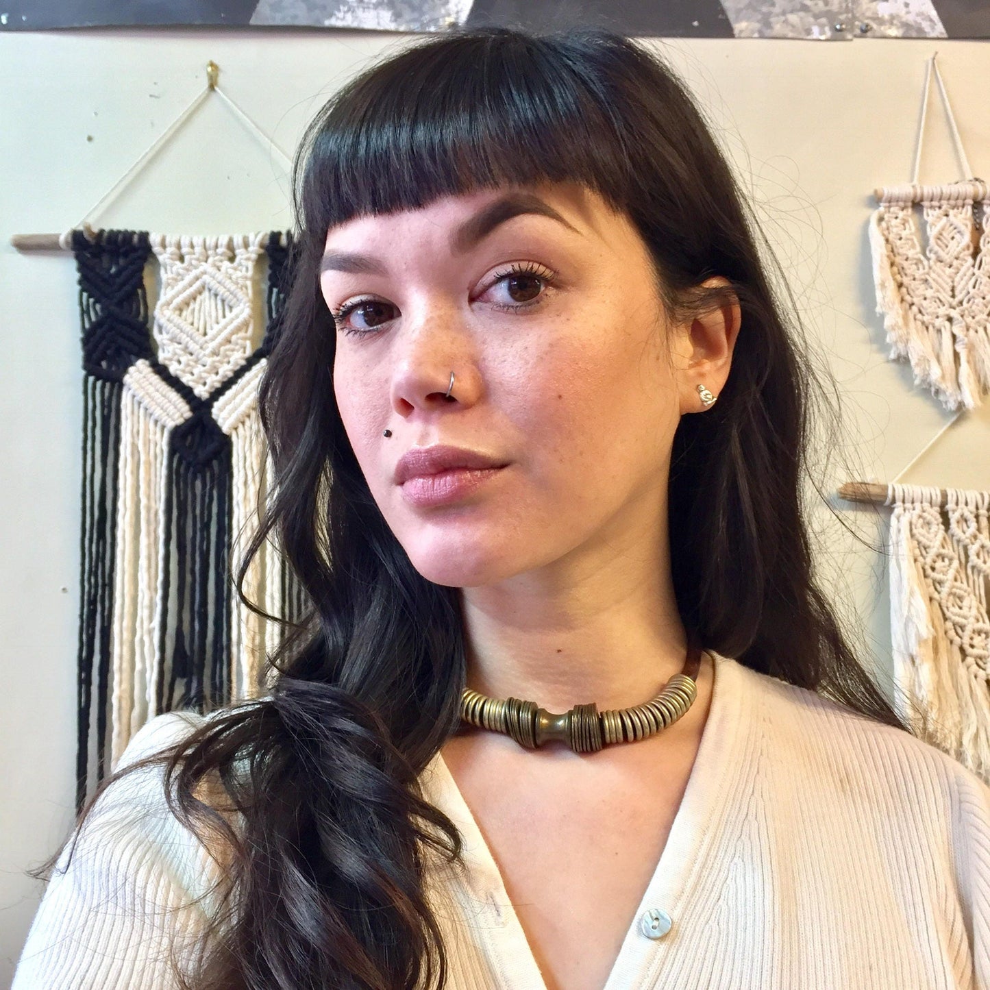 The image shows a young woman with long dark hair, wearing a cream colored sweater or top, and looking thoughtfully off to the side. She has a brass colored coiled choker necklace around her neck, which appears to be a vintage or ethnic style statement piece of jewelry. The background contains some macrame or woven wall hangings.