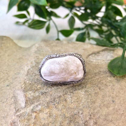 Vintage sterling silver ring with large, hammered rope design setting a pale pink quartz stone, displayed on natural stone surface with greenery in soft focus background.