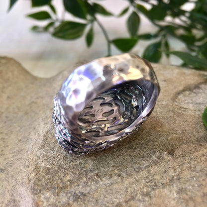 Vintage sterling silver ring with a large, iridescent quartz stone in a hammered, rope-style setting, placed on a textured stone surface with greenery in the background.