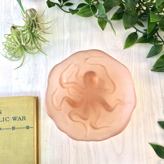 Vintage peach octopus glass dish on a wooden surface with green plants and an old book beside it, perfect for jewelry or as a decorative catch-all.