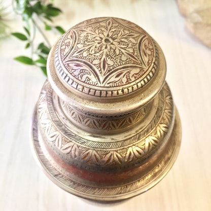 Vintage etched metal cocktail shaker with intricate floral design in silver and red tones, stacked on marble surface with greenery in background, mixology barware from India.