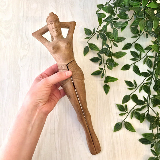 Vintage wooden hand-carved nutcracker figurine shaped like a nude woman, displayed next to greenery against a light wooden background. Primitive novelty home decor piece for serving or cracking nuts.