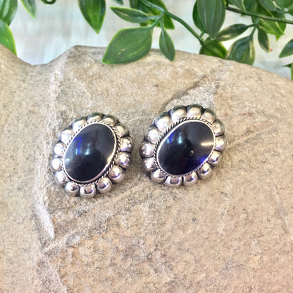 Vintage sterling silver clip-on earrings with black onyx stones in a Southwestern style, featuring large oval cabochon gemstones set in decorative silver bezels, displayed on a stone surface with greenery in the background.